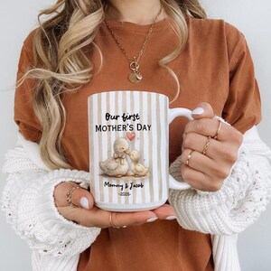 May include: White ceramic mug with vertical beige stripes. The mug features the text "Our first MOTHER'S DAY" and a watercolor illustration of two ducks. Below the illustration is the text "Mommy & Jacob -2026".