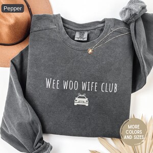 May include: A dark gray sweatshirt with the text "Wee Woo Wife Club" and a police car graphic in white. The sweatshirt has a crew neck and long sleeves. A brown hat is visible in the upper left corner.