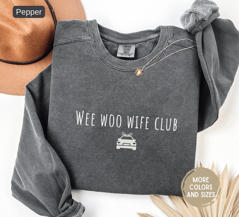 May include: A dark gray sweatshirt with the text "Wee Woo Wife Club" and a police car graphic in white. The sweatshirt has a crew neck and long sleeves. A brown hat is visible in the upper left corner.