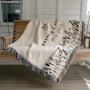 May include: Cream-colored woven throw blanket with black fringe, draped over a wooden swing. The blanket features a heart and the names "AMANDA & CONA" with the date "06-23-2025" in black script. Black floral design along the edge.