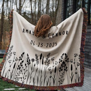 May include: Cream-colored woven throw blanket with black floral design and fringed edges. The blanket features the names "AMAN & CONNOR" and the date "06-23-2025" in black lettering. The fringe has a mix of colors.