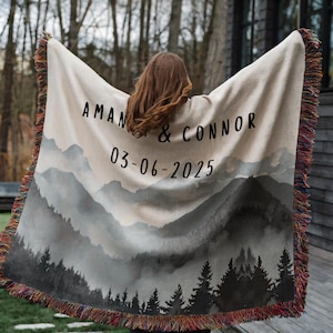 May include: A beige woven blanket with a mountain landscape design and fringed edges. The blanket features the names "AMAN & CONNOR" and the date "03-06-2025" in black lettering. The design includes a forest silhouette.