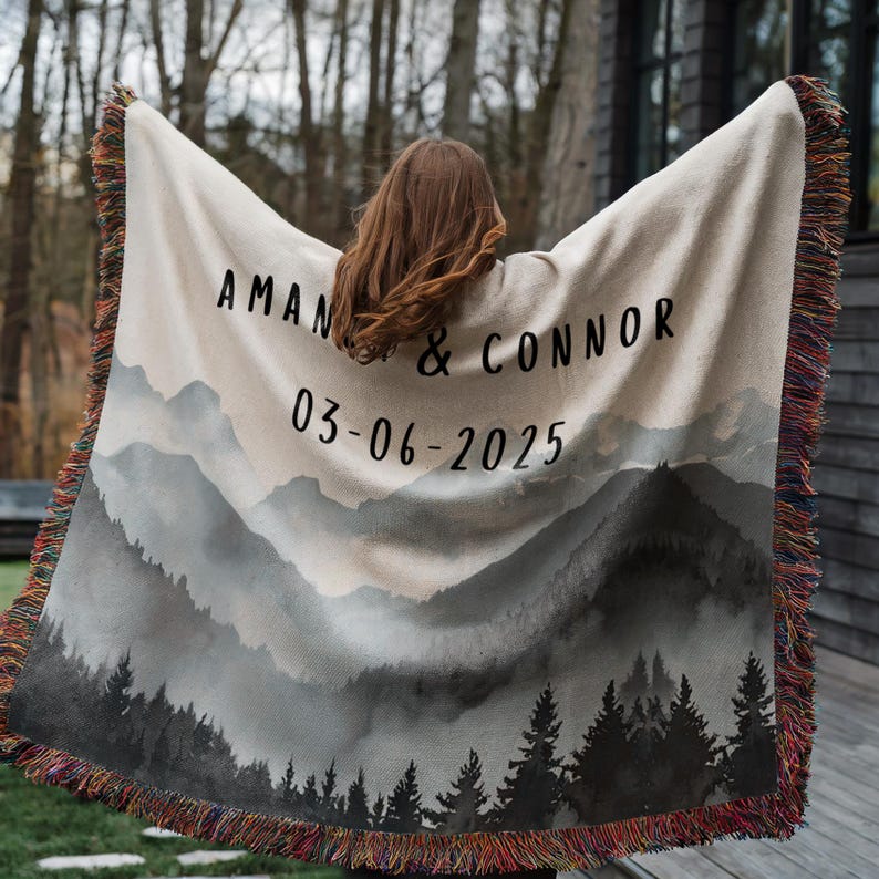 May include: A beige woven blanket with a mountain landscape design and fringed edges. The blanket features the names "AMAN & CONNOR" and the date "03-06-2025" in black lettering. The design includes a forest silhouette.