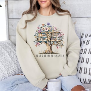 Puede incluir: Una sudadera de color crema con un dise&ntilde;o de acuarela de un &aacute;rbol con libros como ramas y flores coloridas. El texto "JUST ONE MORE CHAPTER" est&aacute; impreso debajo del &aacute;rbol. La sudadera es de estilo casual y c&oacute;modo.