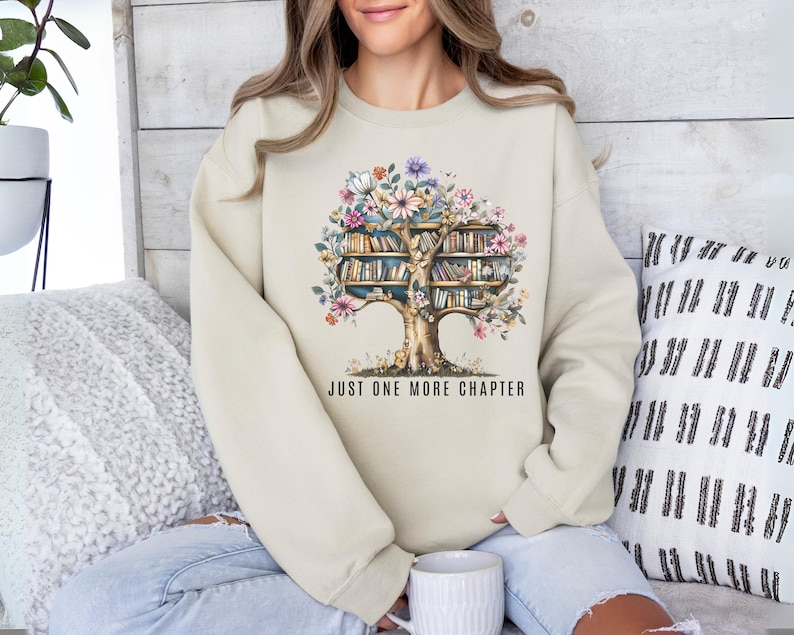 Puede incluir: Una sudadera de color crema con un dise&ntilde;o de acuarela de un &aacute;rbol con libros como ramas y flores coloridas. El texto "JUST ONE MORE CHAPTER" est&aacute; impreso debajo del &aacute;rbol. La sudadera es de estilo casual y c&oacute;modo.