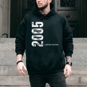 Pu&ograve; includere: Felpa con cappuccio nera con l'anno "2005" stampato verticalmente in bianco, insieme al testo "LIMITED EDITION". La felpa ha una tasca frontale e un cappuccio. La persona indossa un orologio nero e un braccialetto nero.