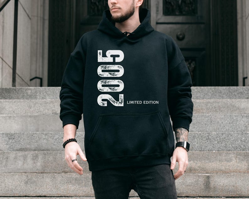 Pu&ograve; includere: Felpa con cappuccio nera con l'anno "2005" stampato verticalmente in bianco, insieme al testo "LIMITED EDITION". La felpa ha una tasca frontale e un cappuccio. La persona indossa un orologio nero e un braccialetto nero.