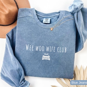 May include: A blue crew neck sweatshirt with the text "Wee Woo Wife Club" and a police car graphic. The sweatshirt is a faded blue color and has long sleeves. A gold necklace is draped across the front.