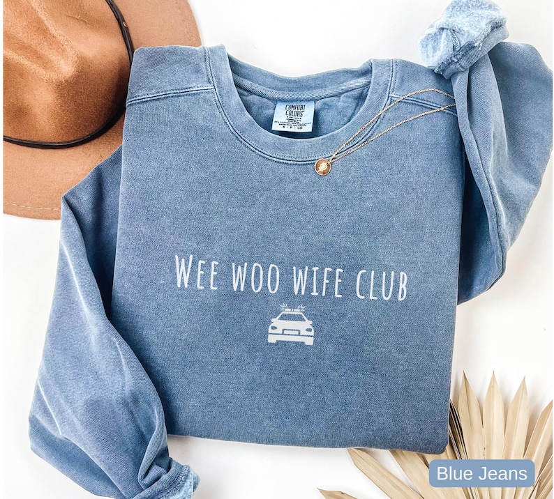 May include: A blue crew neck sweatshirt with the text "Wee Woo Wife Club" and a police car graphic. The sweatshirt is a faded blue color and has long sleeves. A gold necklace is draped across the front.
