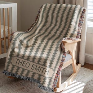 May include: A beige and green striped throw blanket with the name "THEO SMITH" printed on it, draped over a wooden rocking chair. The blanket has a fringed edge with multiple colors.
