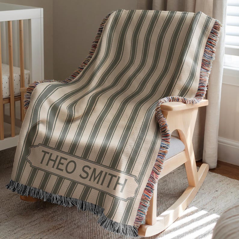 May include: A beige and green striped throw blanket with the name "THEO SMITH" printed on it, draped over a wooden rocking chair. The blanket has a fringed edge with multiple colors.