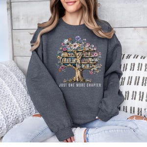 Puede incluir: Una sudadera gris oscuro con un dise&ntilde;o de &aacute;rbol cuyas hojas son libros y flores. El texto "JUST ONE MORE CHAPTER" est&aacute; impreso debajo. La sudadera la lleva una persona.