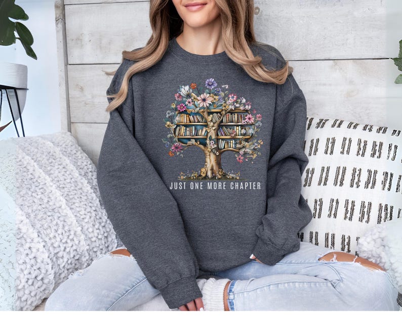 Puede incluir: Una sudadera gris oscuro con un dise&ntilde;o de &aacute;rbol cuyas hojas son libros y flores. El texto "JUST ONE MORE CHAPTER" est&aacute; impreso debajo. La sudadera la lleva una persona.