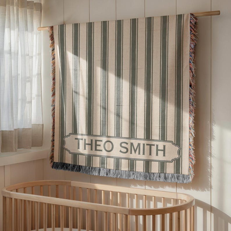 May include: A beige and gray striped blanket with the name "THEO SMITH" displayed in a gray banner. The blanket is hanging on a wooden rod, with a wooden crib visible below. The blanket has fringed edges.