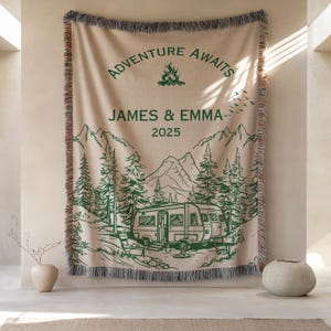 May include: Beige woven throw blanket with green illustrations of a camper van, mountains, and trees. The text "ADVENTURE AWAITS" and "JAMES & EMMA 2025" are printed in green. The blanket has a fringed edge.