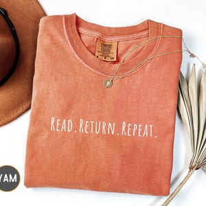 May include: An orange Comfort Colors t-shirt with the words "READ. RETURN. REPEAT." in white. A gold necklace with a pendant rests on the shirt. A brown hat and dried palm leaves are in the background.