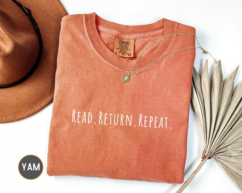 May include: An orange Comfort Colors t-shirt with the words "READ. RETURN. REPEAT." in white. A gold necklace with a pendant rests on the shirt. A brown hat and dried palm leaves are in the background.