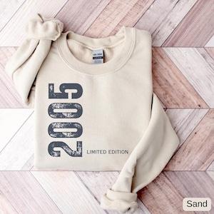 Pu&ograve; includere: Felpa color sabbia con l'anno "2005" stampato verticalmente in grigio scuro, insieme alle parole "Limited Edition". L'etichetta del marchio Gildan &egrave; visibile. La felpa &egrave; piegata con le maniche arrotolate.