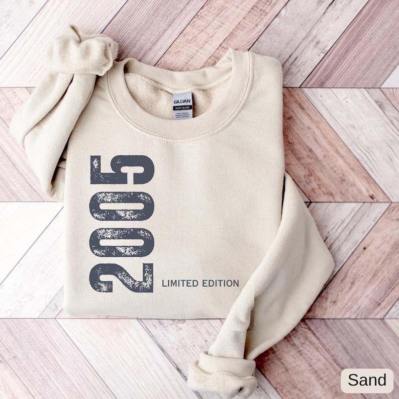 Pu&ograve; includere: Felpa color sabbia con l'anno "2005" stampato verticalmente in grigio scuro, insieme alle parole "Limited Edition". L'etichetta del marchio Gildan &egrave; visibile. La felpa &egrave; piegata con le maniche arrotolate.