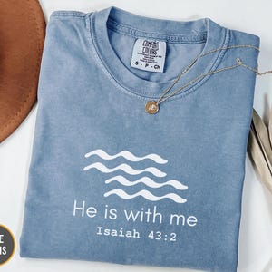 May include: A blue t-shirt with the text "He is with me" and "Isaiah 43:2" in white. The shirt has a crew neck and short sleeves. A gold necklace with a pendant rests on the shirt. A brown hat and dried leaves are in the background.