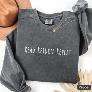 May include: A dark gray sweatshirt with the words "READ. RETURN. REPEAT." in white lettering. The sweatshirt has a crew neck and long sleeves. A gold necklace is draped on the front. A brown hat is in the background.