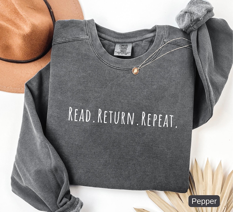May include: A dark gray sweatshirt with the words "READ. RETURN. REPEAT." in white lettering. The sweatshirt has a crew neck and long sleeves. A gold necklace is draped on the front. A brown hat is in the background.