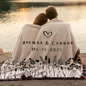 May include: A cream-colored blanket with a black floral design along the bottom edge. The blanket features a heart symbol and the names "AMANDA & CONNOR" with the date "06-23-2025" printed in black. The blanket has a fringed edge.
