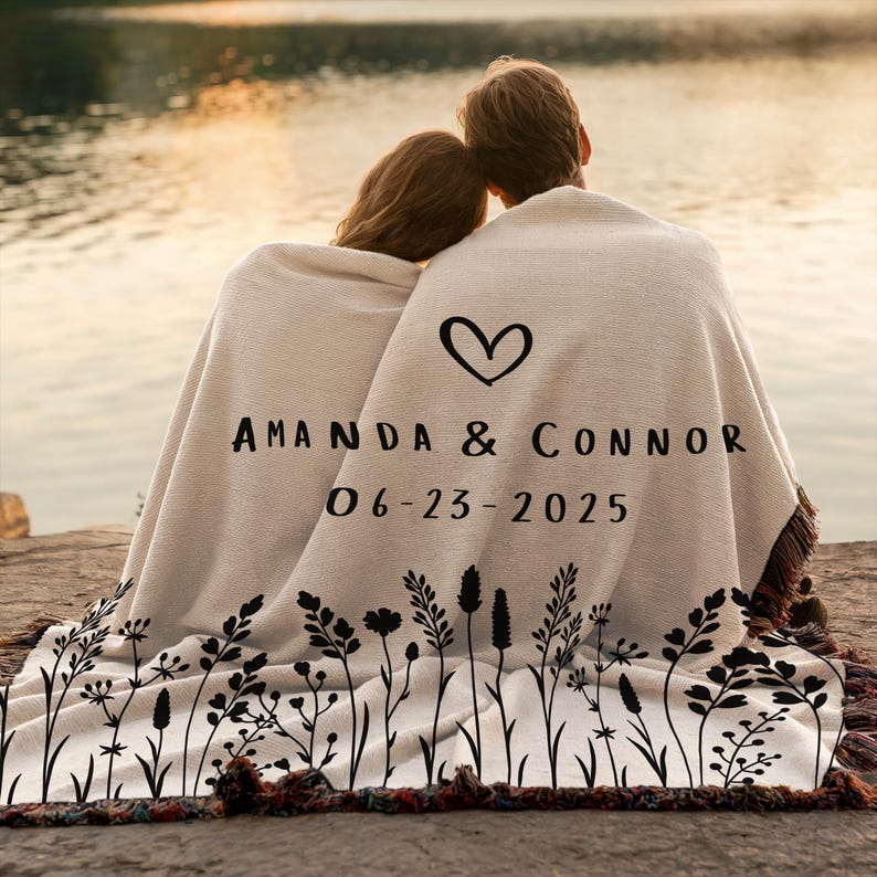May include: A cream-colored blanket with a black floral design along the bottom edge. The blanket features a heart symbol and the names "AMANDA & CONNOR" with the date "06-23-2025" printed in black. The blanket has a fringed edge.