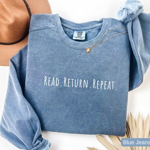 May include: A blue crew neck sweatshirt with the text "READ. RETURN. REPEAT." in white. The sweatshirt has long sleeves and a relaxed fit. A gold necklace with a small pendant is draped across the front. A brown hat is in the background.
