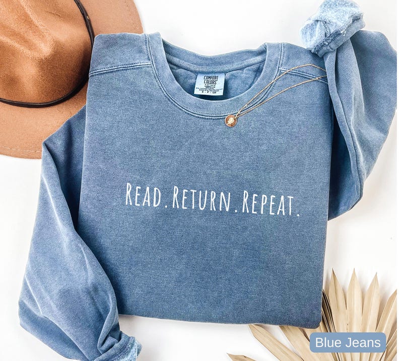 May include: A blue crew neck sweatshirt with the text "READ. RETURN. REPEAT." in white. The sweatshirt has long sleeves and a relaxed fit. A gold necklace with a small pendant is draped across the front. A brown hat is in the background.