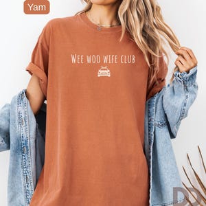 May include: A yam-colored t-shirt with the text "WEE WOO WIFE CLUB" and a car graphic. The shirt has rolled-up sleeves and is paired with a denim jacket. The shirt is a solid color and has a relaxed fit.