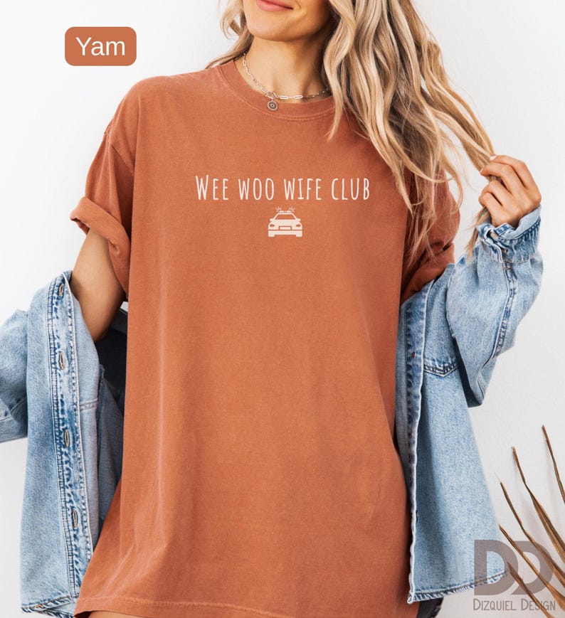 May include: A yam-colored t-shirt with the text "WEE WOO WIFE CLUB" and a car graphic. The shirt has rolled-up sleeves and is paired with a denim jacket. The shirt is a solid color and has a relaxed fit.