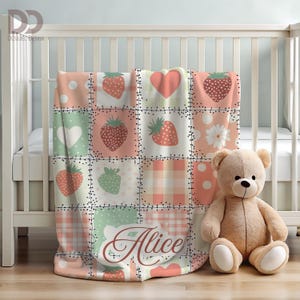 May include: A patchwork baby blanket with strawberry and heart patterns in shades of pink, green, and white. The blanket is draped over a crib and features the name "Alice" in cursive script. A teddy bear sits beside the crib.