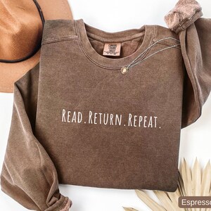 May include: Espresso-colored sweatshirt with the text "READ. RETURN. REPEAT." in white. The crew neck sweatshirt has long sleeves and a relaxed fit. A gold-colored necklace is draped across the front. A brown hat is visible in the upper left corner.