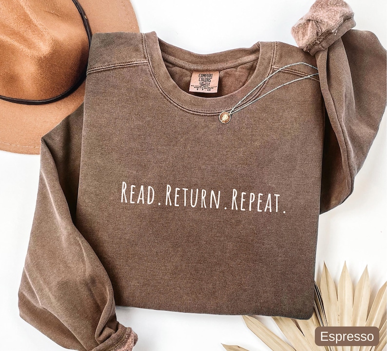 May include: Espresso-colored sweatshirt with the text "READ. RETURN. REPEAT." in white. The crew neck sweatshirt has long sleeves and a relaxed fit. A gold-colored necklace is draped across the front. A brown hat is visible in the upper left corner.