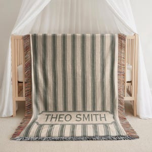 May include: A woven baby blanket with vertical stripes in gray and cream, draped over a crib. The blanket features a rectangular label with the name "THEO SMITH" in gray lettering. The edges have a fringed trim.