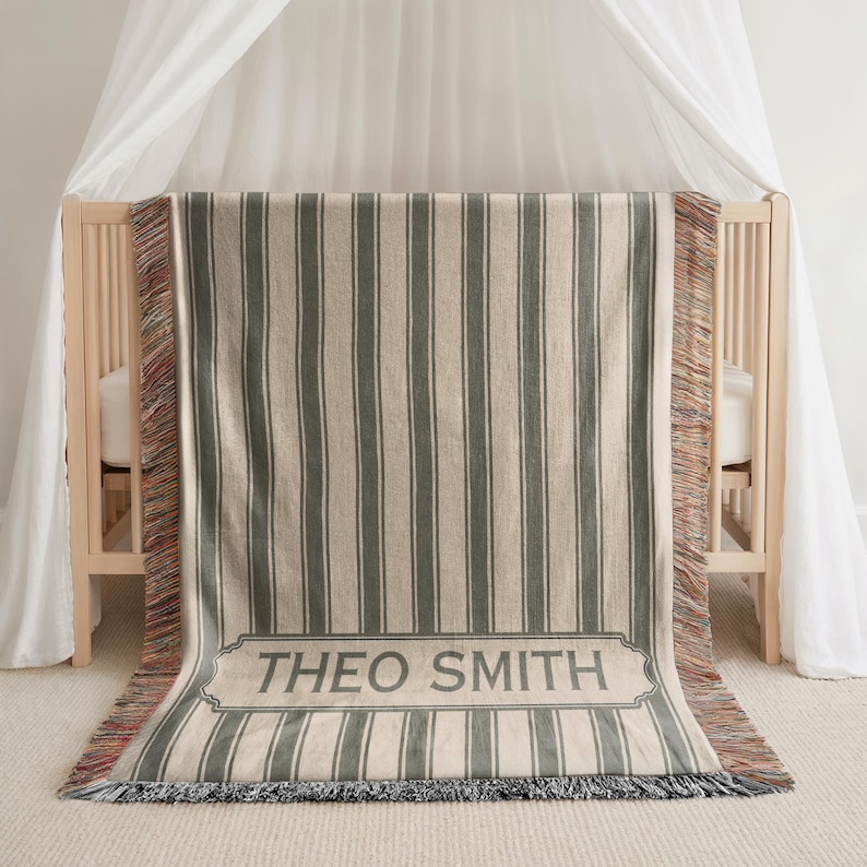 May include: A woven baby blanket with vertical stripes in gray and cream, draped over a crib. The blanket features a rectangular label with the name "THEO SMITH" in gray lettering. The edges have a fringed trim.