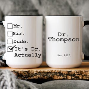 May include: Two white ceramic mugs with black handles and black trim. The mug on the left has a checklist with the options "Mr.", "Sir.", "Dude.", and "It's Dr. Actually" checked. The mug on the right has the text "Dr. Thompson Est. 2025".