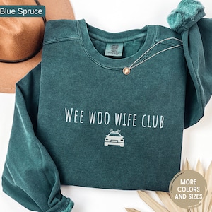 May include: A blue spruce crewneck sweatshirt with the text "WEE WOO WIFE CLUB" and a police car graphic. A gold necklace with a pendant and a green scrunchie are also visible. The sweatshirt is a Comfort Colors brand.