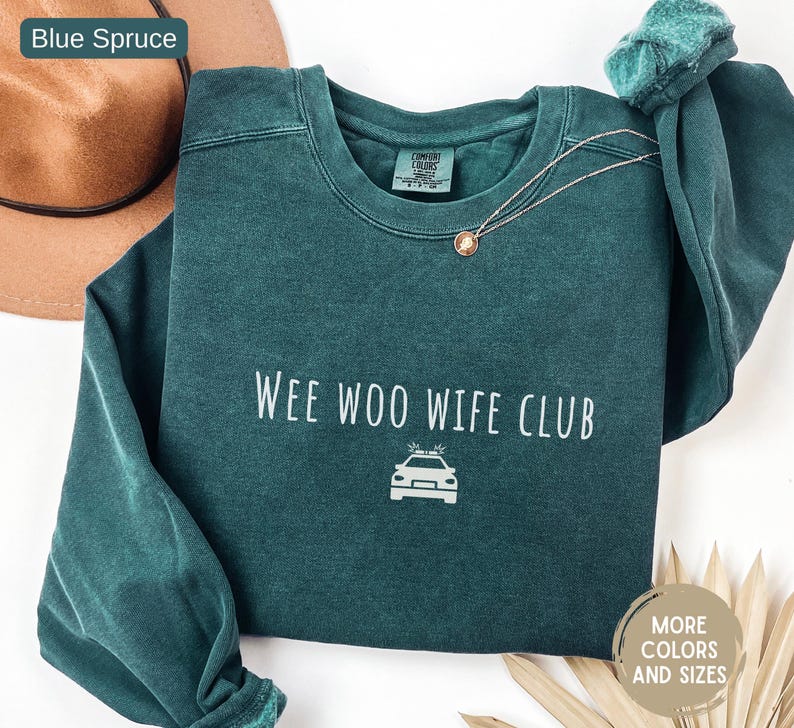 May include: A blue spruce crewneck sweatshirt with the text "WEE WOO WIFE CLUB" and a police car graphic. A gold necklace with a pendant and a green scrunchie are also visible. The sweatshirt is a Comfort Colors brand.