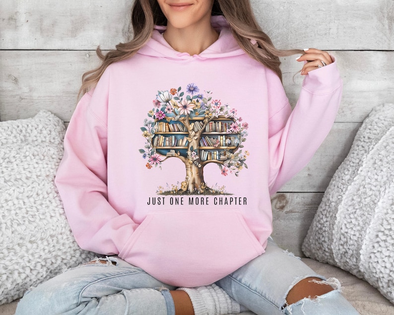 Puede incluir: Sudadera con capucha rosa claro con un dise&ntilde;o de &aacute;rbol con libros y flores coloridas. El texto "JUST ONE MORE CHAPTER" est&aacute; impreso debajo. La sudadera tiene un bolsillo delantero y una capucha.