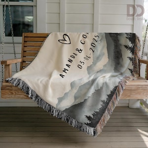 May include: Cream-colored woven throw blanket with a mountain and forest design. The blanket features the names "AMANDA & CO" and the date "03-06-2025" with a heart symbol. The blanket has a fringed edge and is draped over a wooden swing.