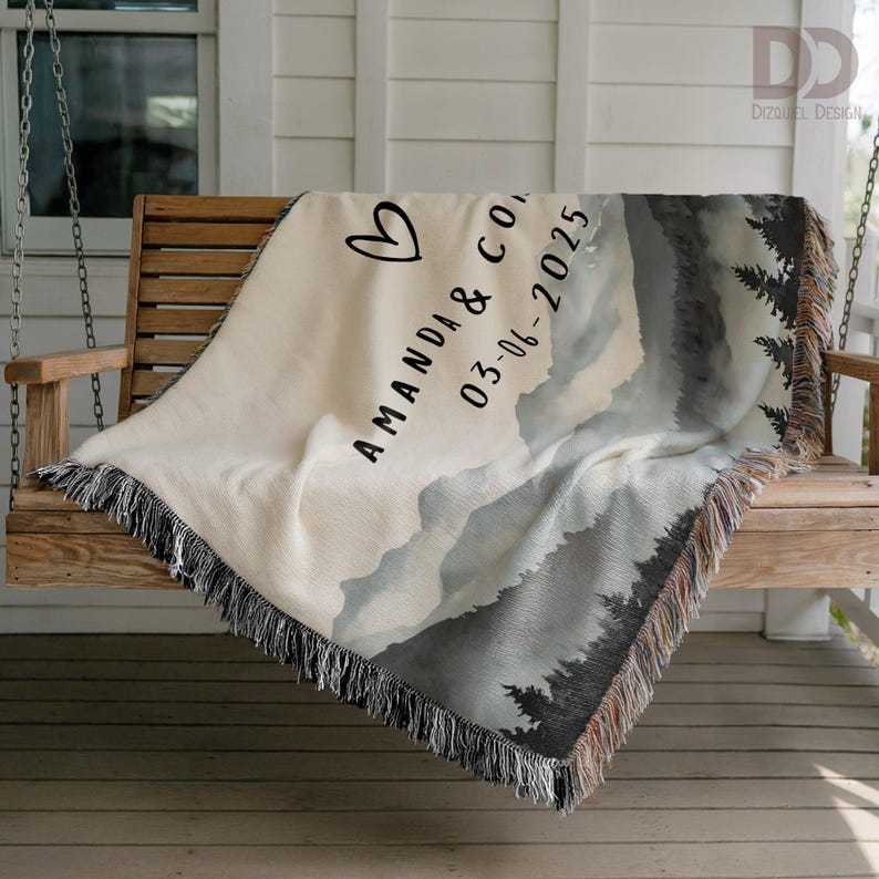 May include: Cream-colored woven throw blanket with a mountain and forest design. The blanket features the names "AMANDA & CO" and the date "03-06-2025" with a heart symbol. The blanket has a fringed edge and is draped over a wooden swing.