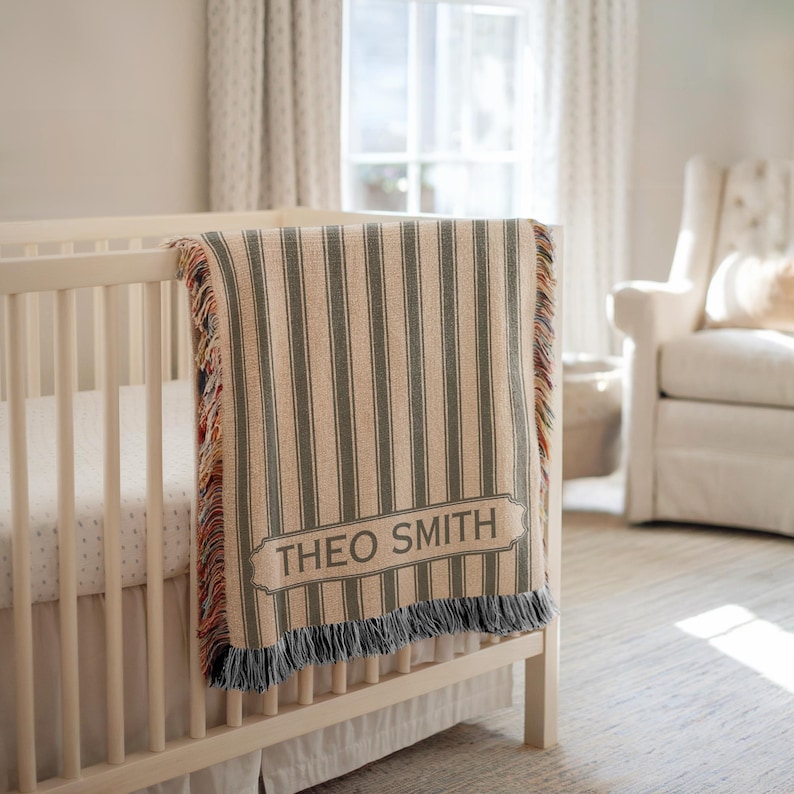 May include: A beige and gray striped baby blanket with the name "THEO SMITH" in a rectangular label is draped over a white crib. The blanket has a fringed edge with a mix of colors. The nursery has a white crib, a white chair, and a window.