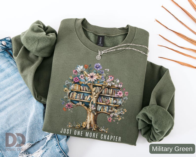 Puede incluir: Sudadera verde militar con un dise&ntilde;o de &aacute;rbol floral y estanter&iacute;as. El texto "JUST ONE MORE CHAPTER" est&aacute; impreso debajo del &aacute;rbol. Un collar plateado est&aacute; sobre la sudadera. La sudadera est&aacute; doblada junto a unos vaqueros azules.