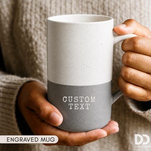 May include: A tall, two-toned ceramic mug featuring a speckled off-white upper section and a solid grey lower section. The grey portion is engraved with the words CUSTOM TEXT in a serif font. The mug has a smooth, cylindrical body and a side handle.
