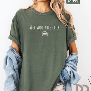 May include: Moss green t-shirt with rolled sleeves. The shirt features the text "WEE WOO WIFE CLUB" above a car graphic. A light blue denim jacket is draped over the shoulders. The shirt is from Dizquiel Design.