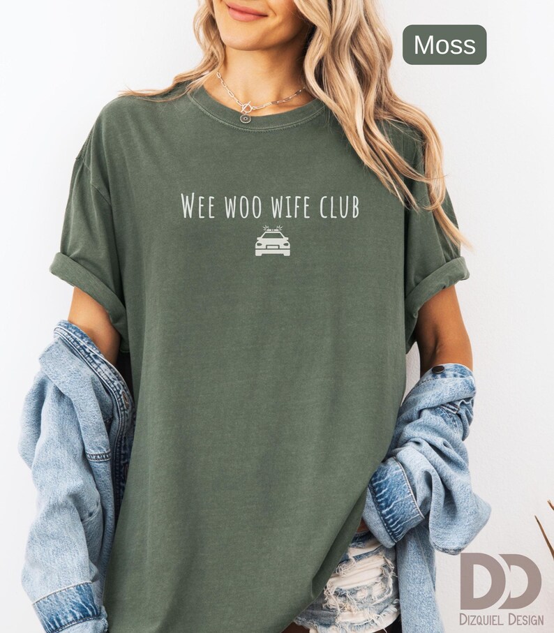 May include: Moss green t-shirt with rolled sleeves. The shirt features the text "WEE WOO WIFE CLUB" above a car graphic. A light blue denim jacket is draped over the shoulders. The shirt is from Dizquiel Design.