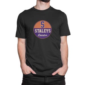 May include: Black t-shirt featuring a circular design with the word "STALEYS" and "Decatur" in white text. The design includes an orange and purple color scheme with the letter "S" in orange.
