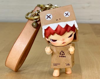 May include: A brown cardboard box character keychain with a red mouth, white teeth, and a brown leather strap. The box has a red X on each side and the text "Dangerous Cargo" and "HRN" printed on the front.